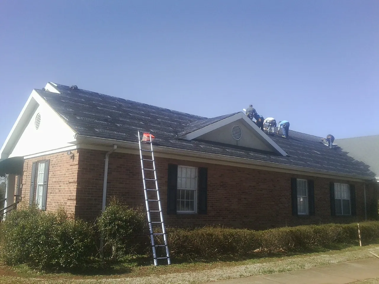 Crew tearing shingles off a brick ranch roof with a ladder set — Emergency Roof Repair work by our crew in Tallassee