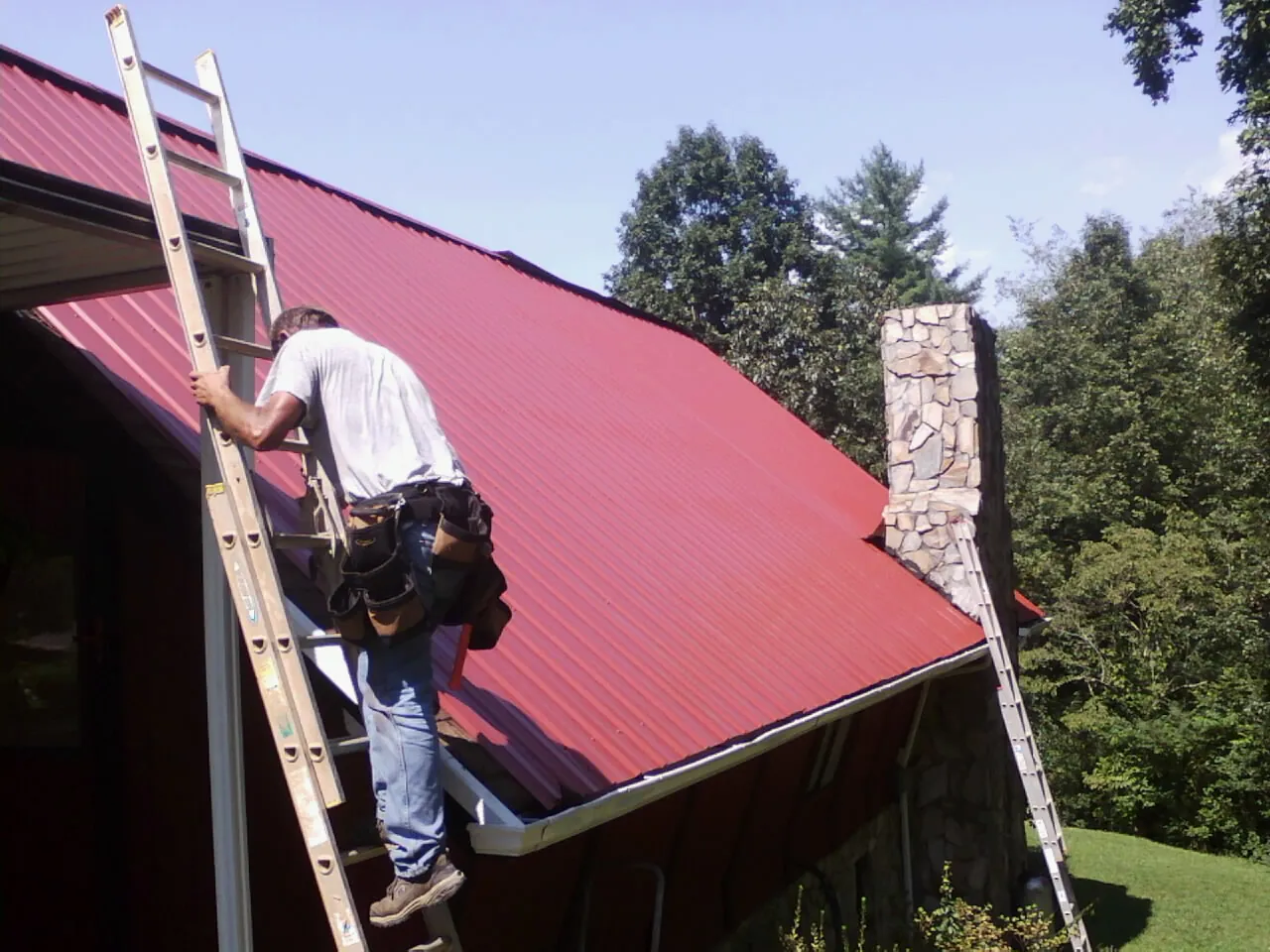 Roofer on a ladder beside a red metal roof and stone chimney — Soffit & Fascia Repair by Roofing Brothers in Tallassee